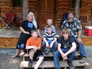 Ron and Caryl in front of a rustic cabin of grandparents with four stair-stepped grandsons.